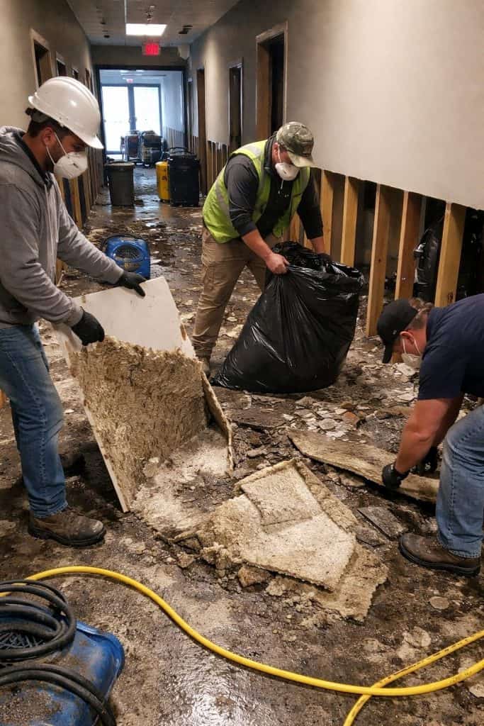 Restoration technicians removing wet drywall and silt-covered debris from a flooded hallway